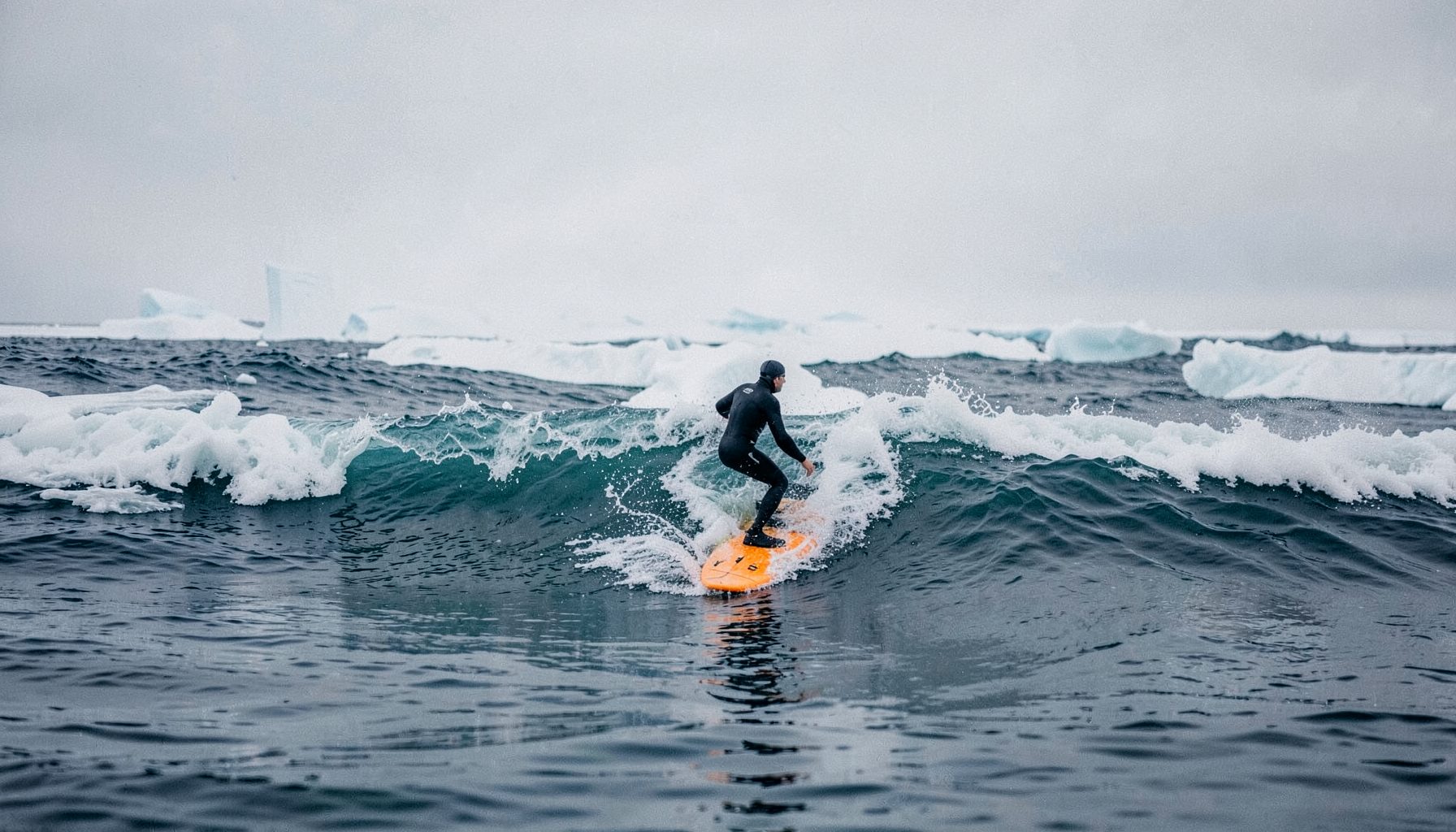 A lone surfer in a thick wetsuit riding a wave on Lake Superior, surrounded by ice formations and a steel-gray winter sky