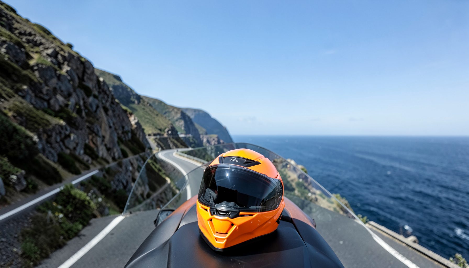 A motorcycle helmet resting on a seat with a coastal highway curving along cliffs in the background under blue skies